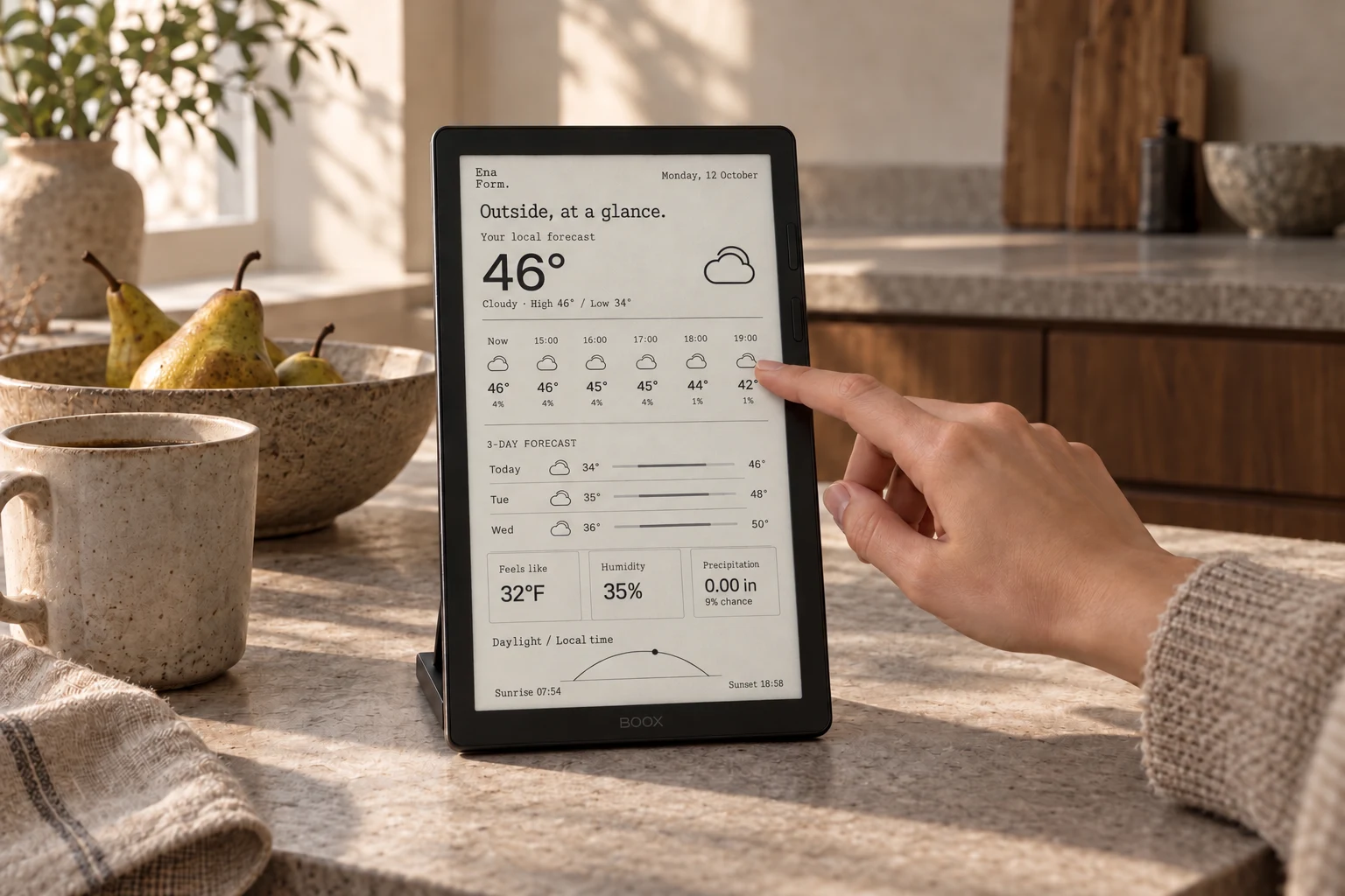 A small e-ink display showing a monochrome weather view beside ceramics on a sunlit kitchen counter, with someone selecting the forecast.