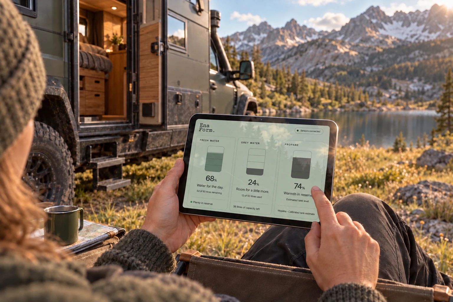 A traveller checking water and propane reserves on an iPad beside a rugged expedition truck at a mountain campsite.